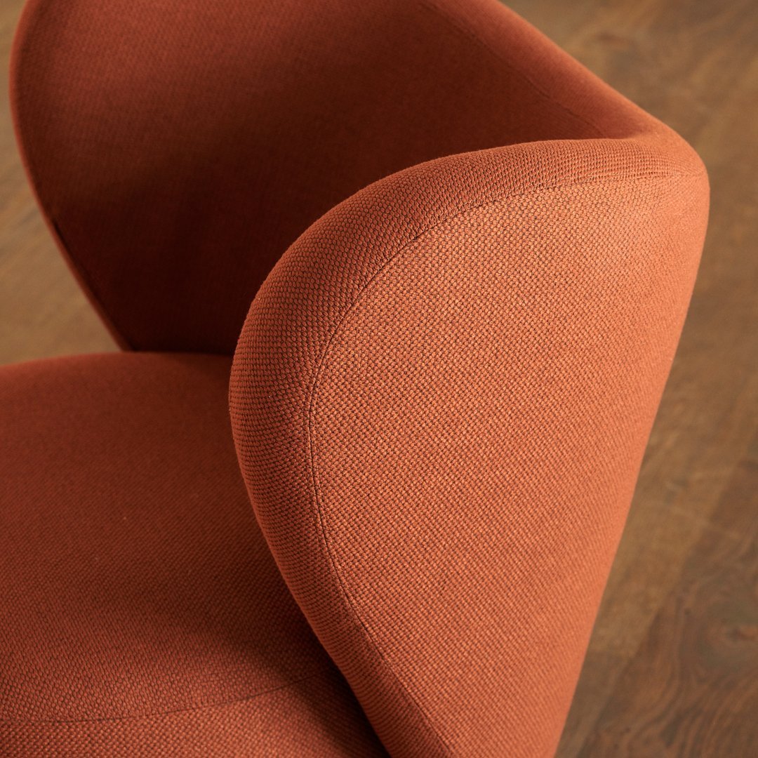 Rust-colored upholstered chair with a smooth, textured finish and curved armrests against a wooden floor backdrop.
