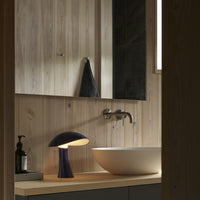 Elegant purple lamp with a smooth, rounded design, set against warm wood paneling and a minimalist black basin.