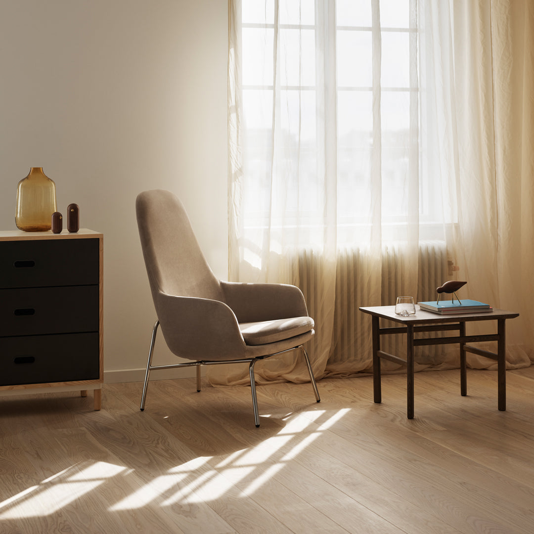 Beige accent chair in a sunlit room with light curtains and wooden flooring, complemented by a black dresser and glass table.