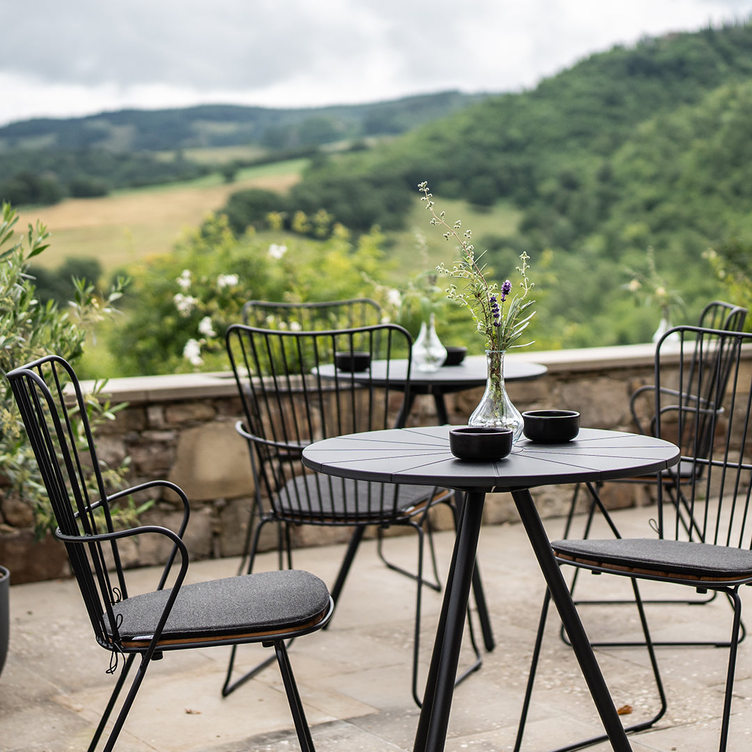 Black metal garden furniture set against a lush green landscape, featuring a round table and dark cushioned chairs.