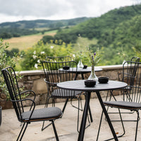 Black metal garden furniture set against a lush green landscape, featuring a round table and dark cushioned chairs.