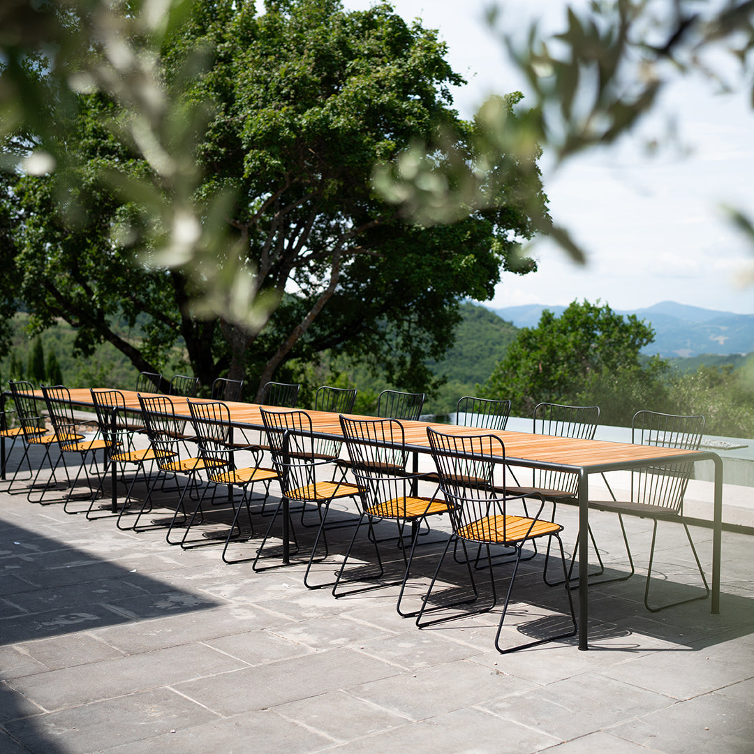 Long black metal dining table with natural wood top and yellow chairs, set against a lush green landscape.