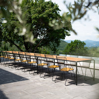 Long black metal dining table with natural wood top and yellow chairs, set against a lush green landscape.