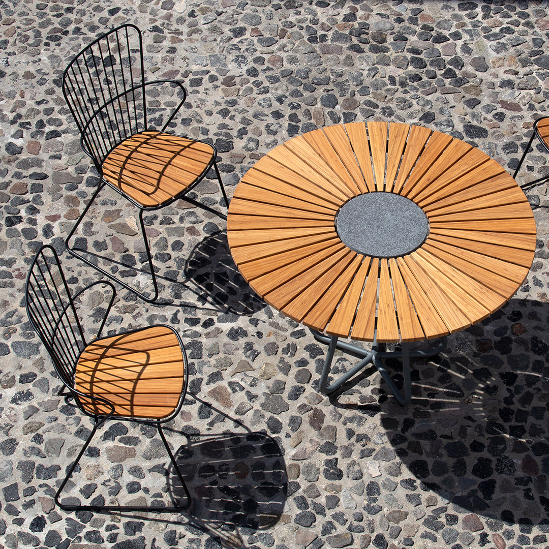 Wooden table with sunburst pattern and black metal chairs on a pebble surface. Warm wood tones contrast with dark metal.