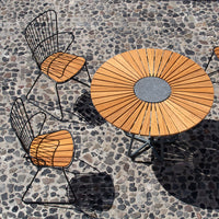 Wooden table with sunburst pattern and black metal chairs on a pebble surface. Warm wood tones contrast with dark metal.