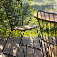 Black wireframe chair on a wooden deck, casting shadows through greenery in a sunlit outdoor setting.