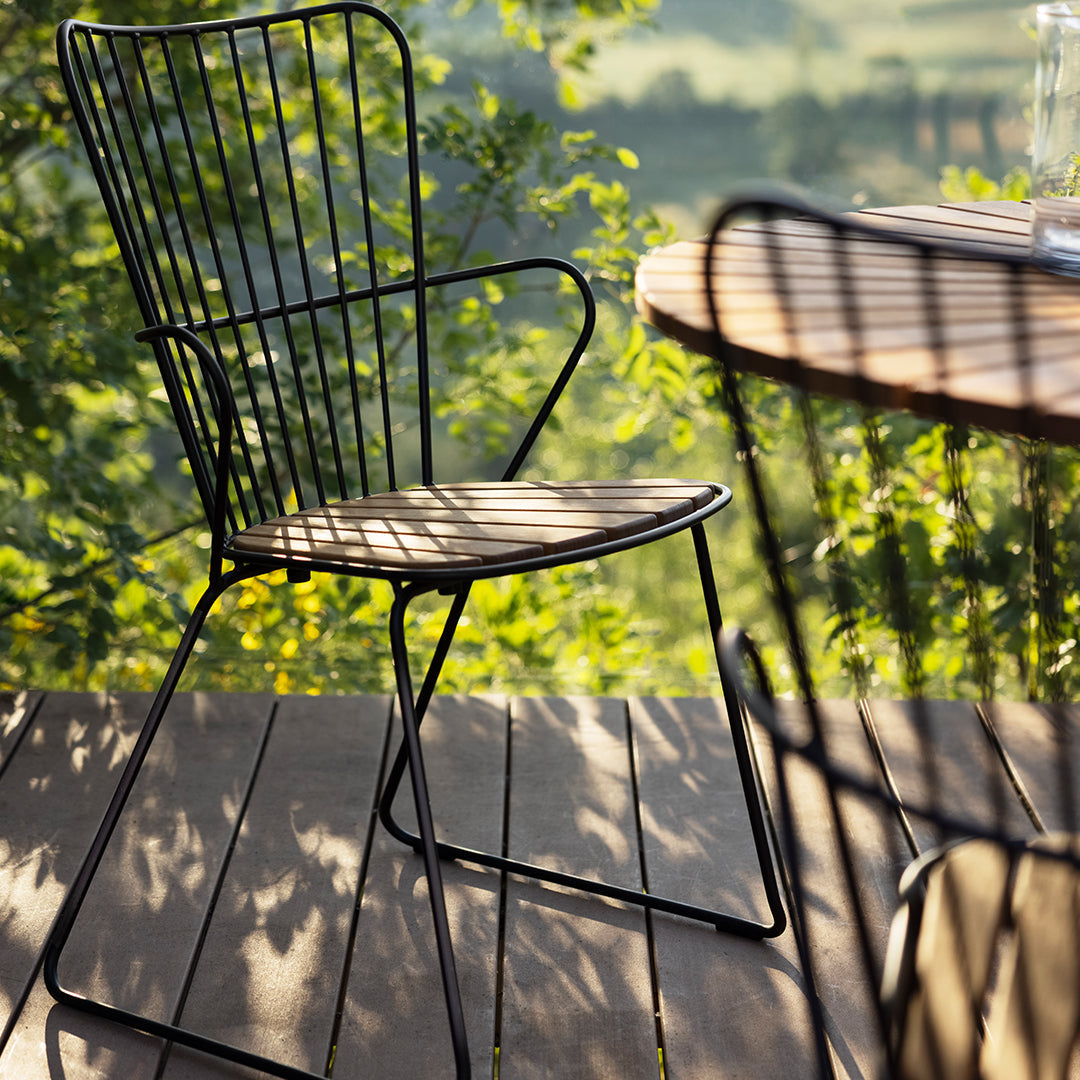 Black wireframe chair on a wooden deck, casting shadows through greenery in a sunlit outdoor setting.