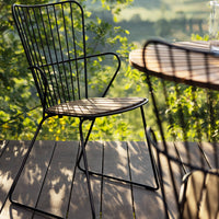 Black wireframe chair on a wooden deck, casting shadows through greenery in a sunlit outdoor setting.