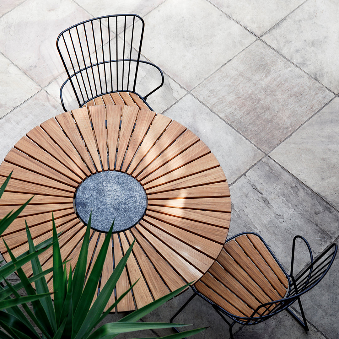Round wooden table with a gray center, paired with black wire chairs against a neutral stone floor, surrounded by greenery.