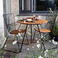 Round wooden table with black metal chairs, set in a natural setting with green grass and white flowers.
