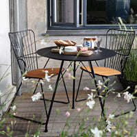 Black circular table with wooden accents and two black wire chairs, set against floral greenery.