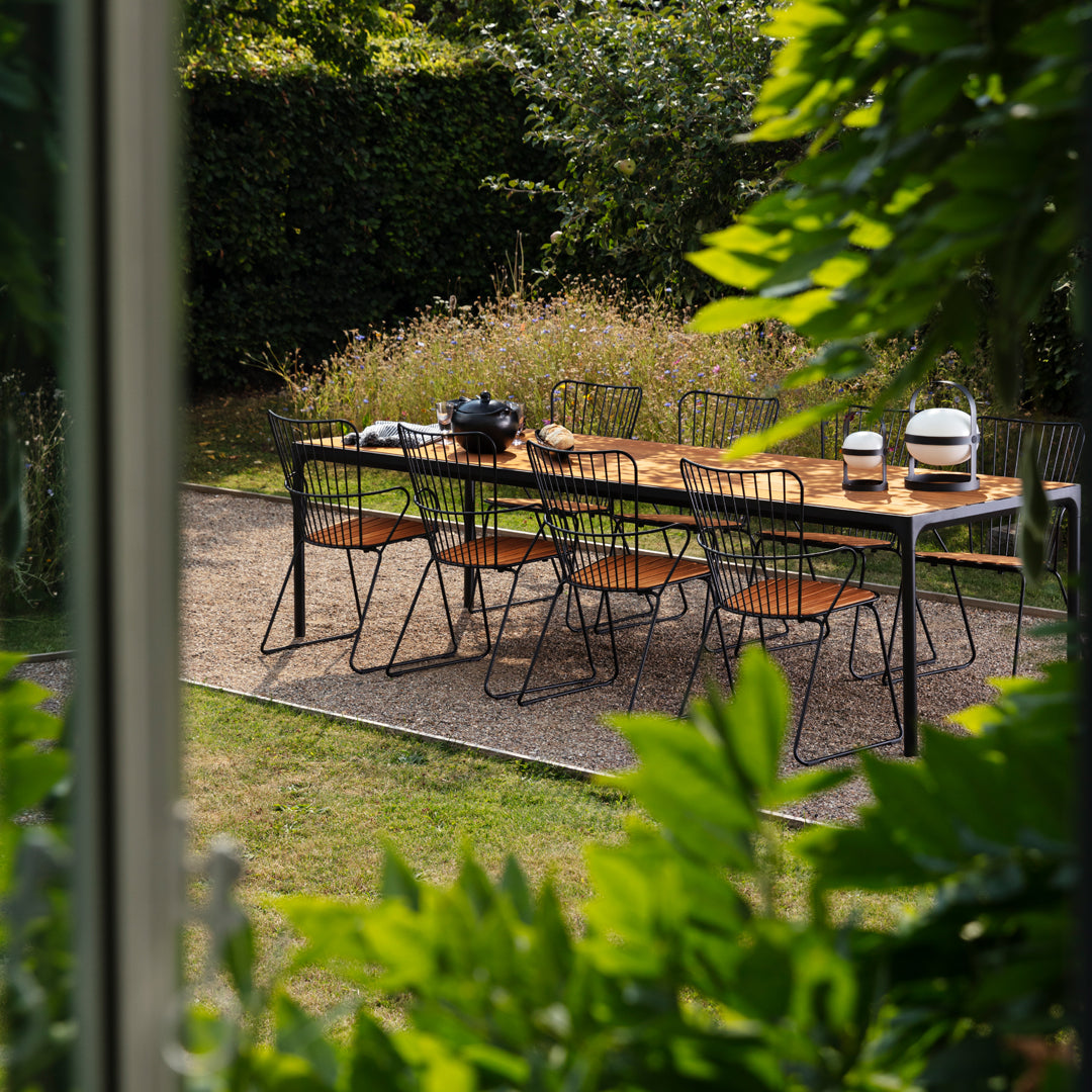 Outdoor dining table with natural wood finish, black metal chairs, and lush green foliage in a sunny garden setting.