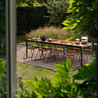 Outdoor dining table with natural wood finish, black metal chairs, and lush green foliage in a sunny garden setting.