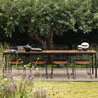 Black metal dining table with wooden accents and matching wire chairs, set against a lush green garden backdrop.