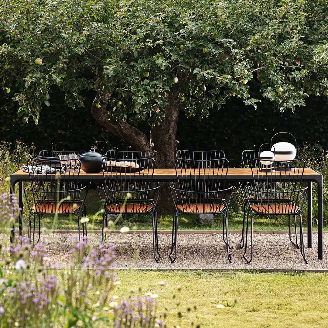 Black metal dining table with wood accents and matching wire chairs, set against a lush green garden backdrop.