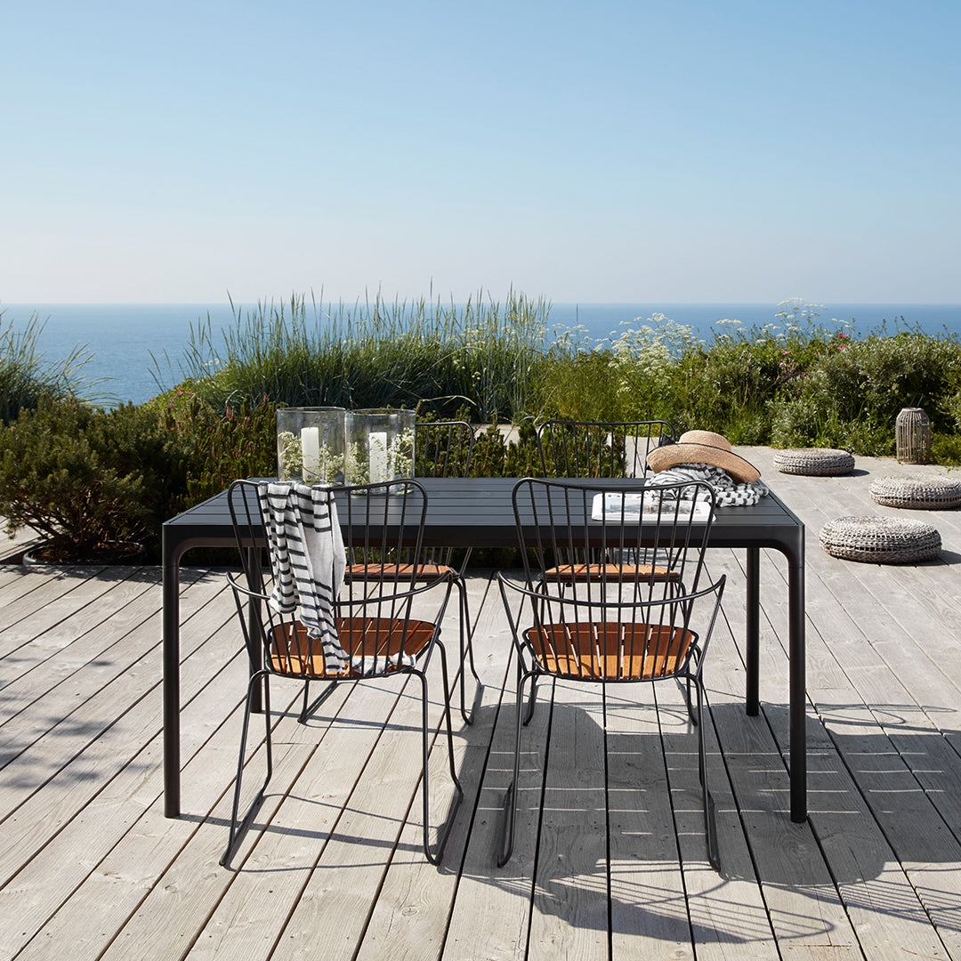 "Black outdoor dining table with striped cushions and wicker chairs, set against a scenic coastal backdrop."
