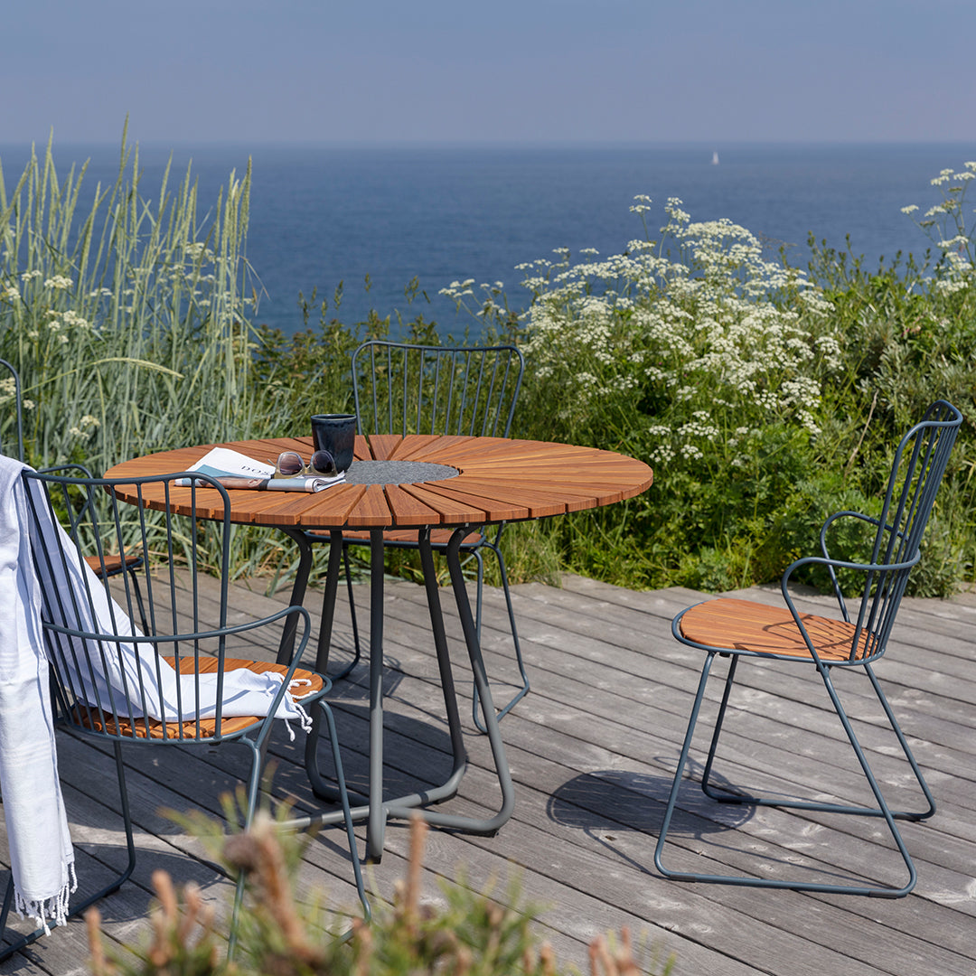 Round wooden table with a natural finish, complemented by blue metal chairs against a coastal backdrop.