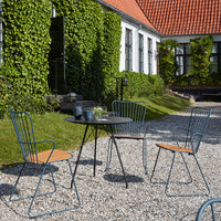 Stylish blue metal chairs and black table with wooden accents, set against lush greenery and a charming red-roofed backdrop.