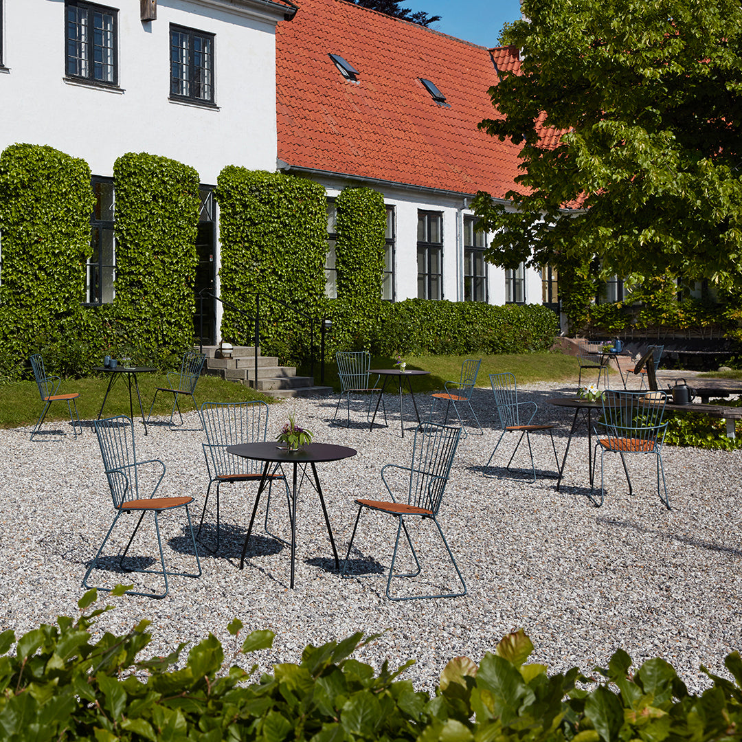 Outdoor patio featuring green hedges, gray gravel, and modern wire chairs with wooden accents against a sunny backdrop.