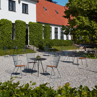 Outdoor patio featuring green hedges, gray gravel, and modern wire chairs with wooden accents against a sunny backdrop.