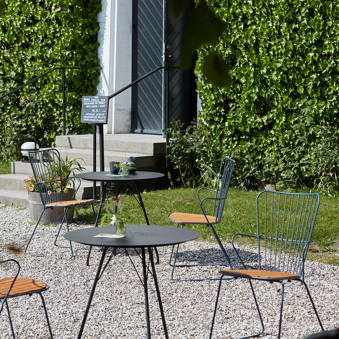 Outdoor seating set featuring gray tables and green wire chairs with wooden accents, surrounded by lush greenery.