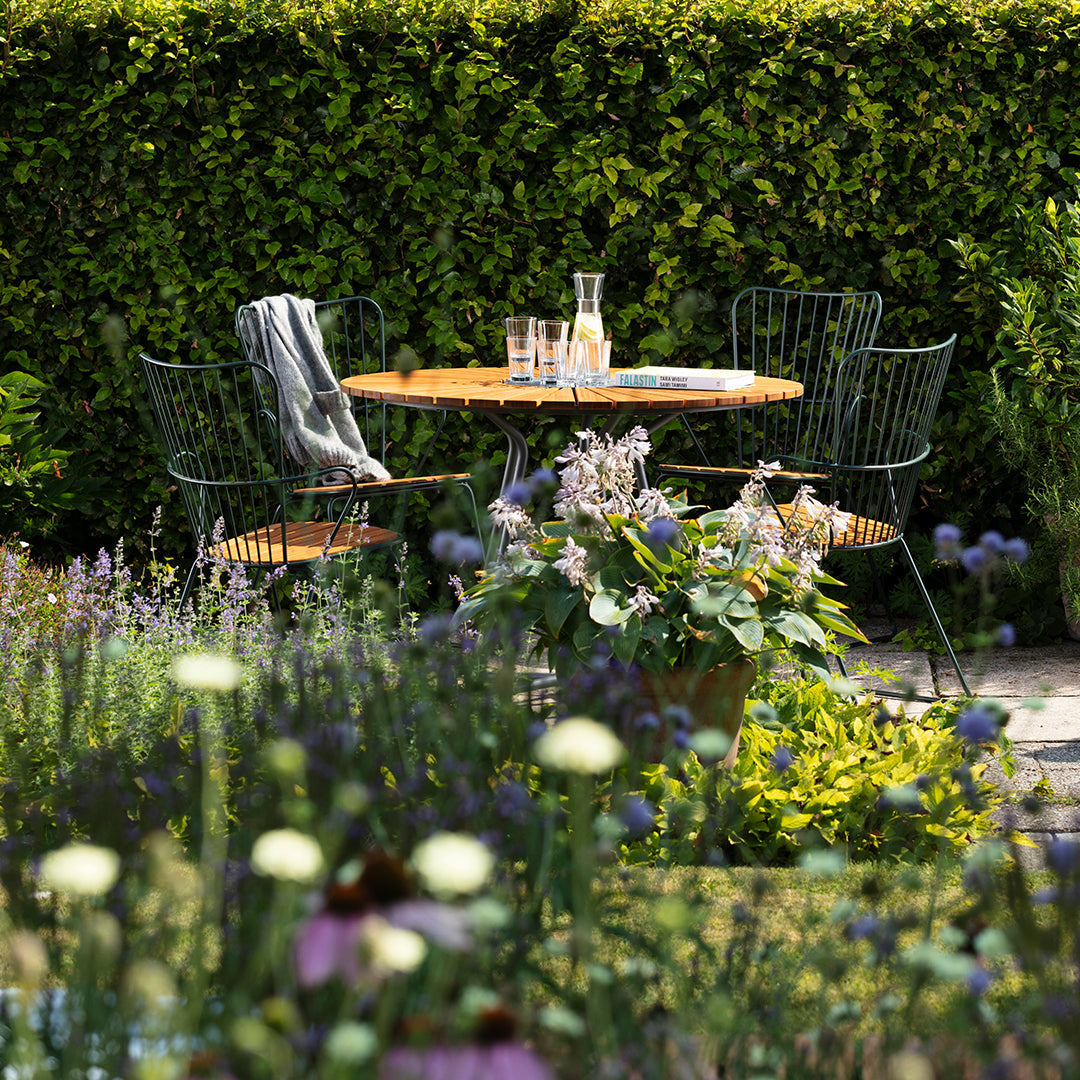 Elegant outdoor dining set with a wooden table, black wire chairs, surrounded by vibrant green foliage and colorful flowers.