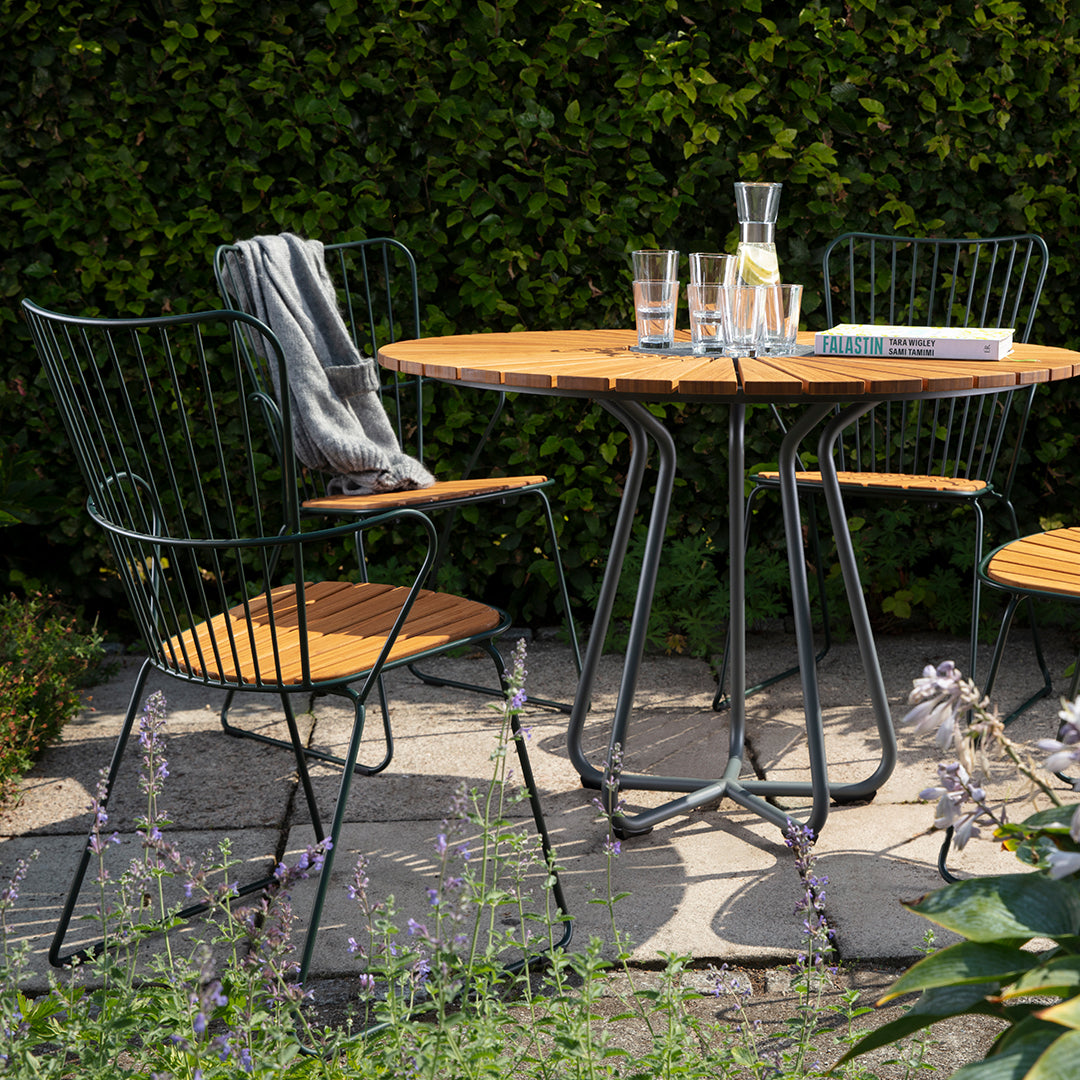 Patio table set featuring a wooden round tabletop, black wire chairs, and a green hedge backdrop. Ideal for outdoor dining.
