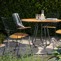 Patio table set featuring a wooden round tabletop, black wire chairs, and a green hedge backdrop. Ideal for outdoor dining.