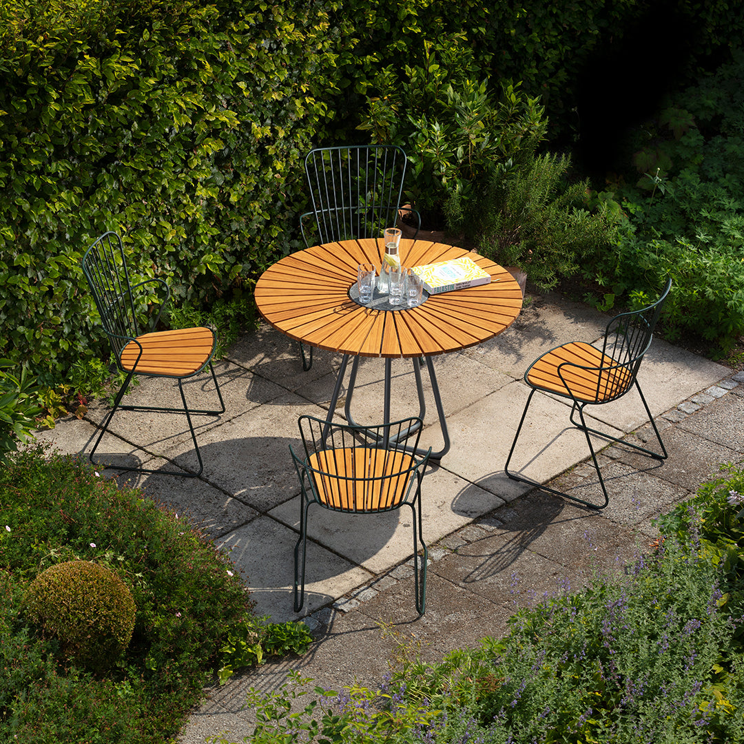 Round wooden patio table with black metal legs, surrounded by black and wooden chairs, set in a lush green garden.