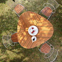 Wooden triangular table with a natural finish, surrounded by metallic chairs with wooden accents. Green grass backdrop.