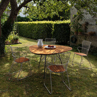"Natural wood round table with minimalistic metal chairs, featuring warm wood tones and sleek silver accents in a green garden."