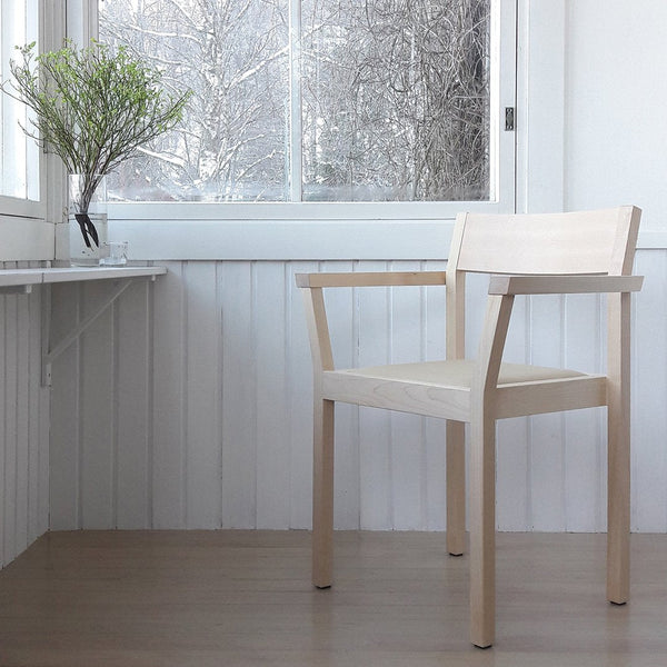 Minimalist beige wooden chair with simple lines, set against a bright, sunlit interior and natural greenery.