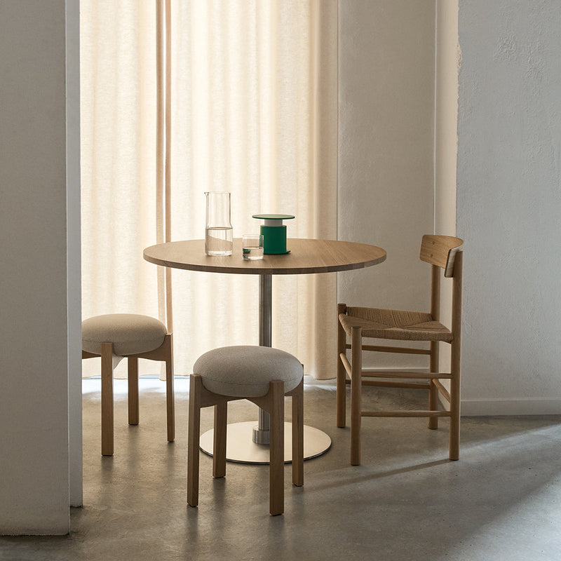 Modern round wooden table with a light beige fabric chair and a green accent piece on a soft grey floor.