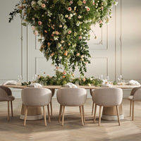 Elegant beige upholstered chairs with light wooden bases, accented by lush green foliage and soft pastel flower arrangements.