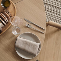 Elegant wooden table setting with neutral-toned plate, linen napkin, clear glassware, and rustic woven basket.