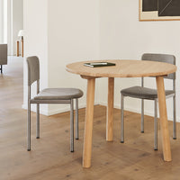 Round wooden table with natural finish, paired with sleek gray chairs on warm wood flooring, modern minimalist design.