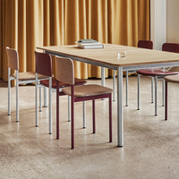 Wooden table with natural finish, surrounded by burgundy and light wood patterned chairs, set against golden curtains.