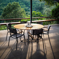Wooden round table with sleek black chairs against a lush green outdoor backdrop, showcasing natural textures and colors.