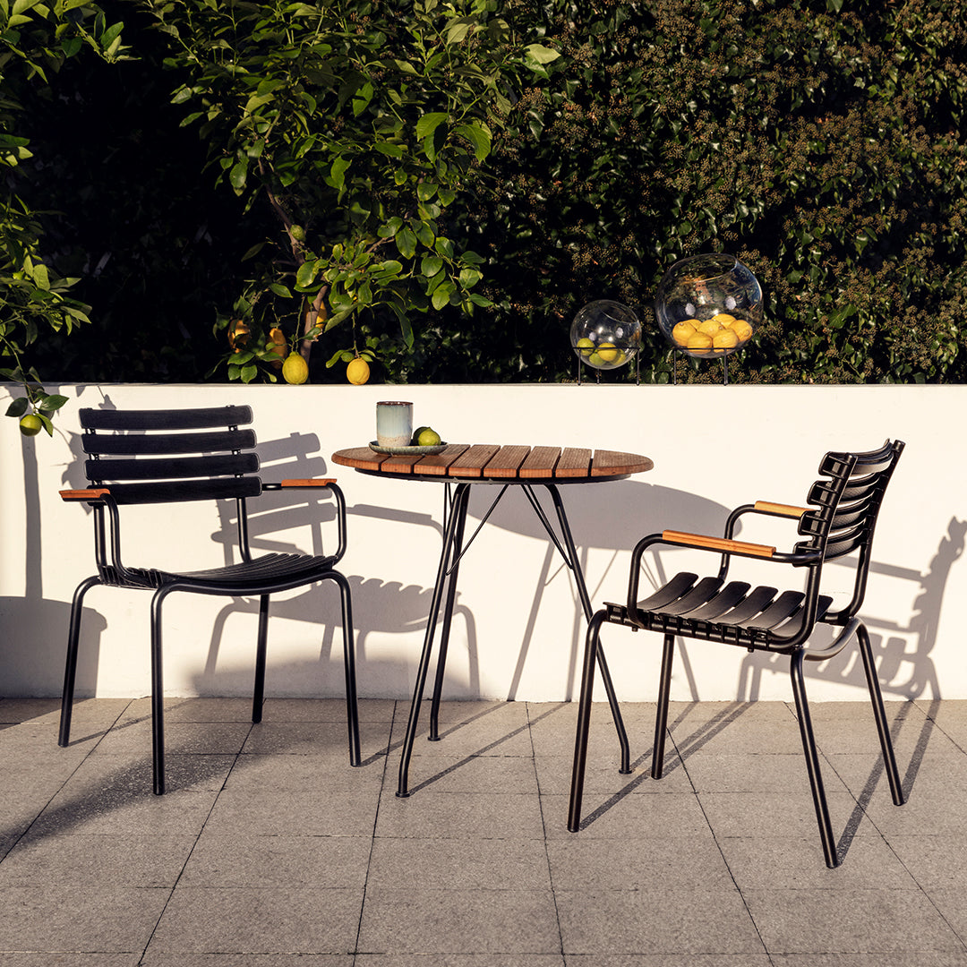 Black metal chairs with wooden accents and a round brown wooden table, set against a lush green backdrop.