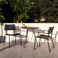 Black metal chairs with wooden accents and a round brown wooden table, set against a lush green backdrop.