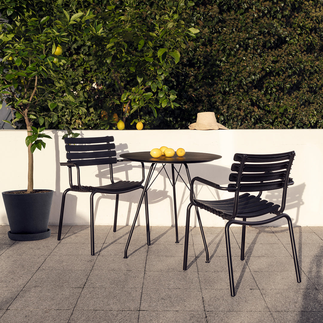 Black metal chairs with horizontal slats and a matching round table on a gray stone patio, surrounded by greenery.