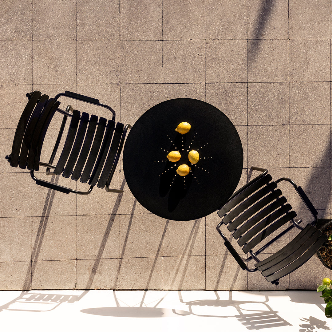 Black outdoor table with four lemons, surrounded by two black slatted chairs on a textured gray tile surface.