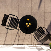 Black outdoor table with four lemons, surrounded by two black slatted chairs on a textured gray tile surface.