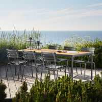 Outdoor dining table with natural wood finish and gray metal chairs, surrounded by greenery and blue ocean backdrop.
