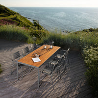 "Rustic wooden dining table with gray metal chairs, set against a scenic ocean backdrop and vibrant greenery."