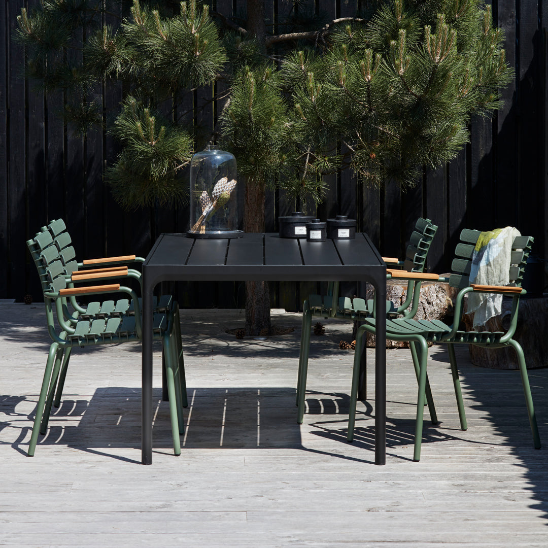 Black outdoor table with green woven chairs, set against a natural backdrop of pine trees and wooden decking.