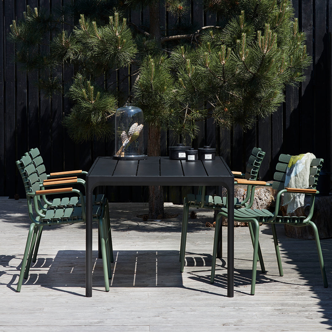Black outdoor dining table with green and wooden accent chairs, set against a dark background of trees and deck.