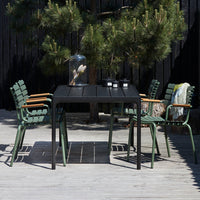 Black outdoor table with green woven chairs, set against a natural backdrop of pine trees and wooden decking.