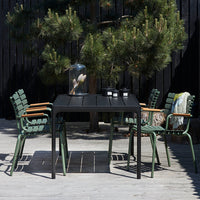 Black outdoor dining table with green and wooden accent chairs, set against a dark background of trees and deck.