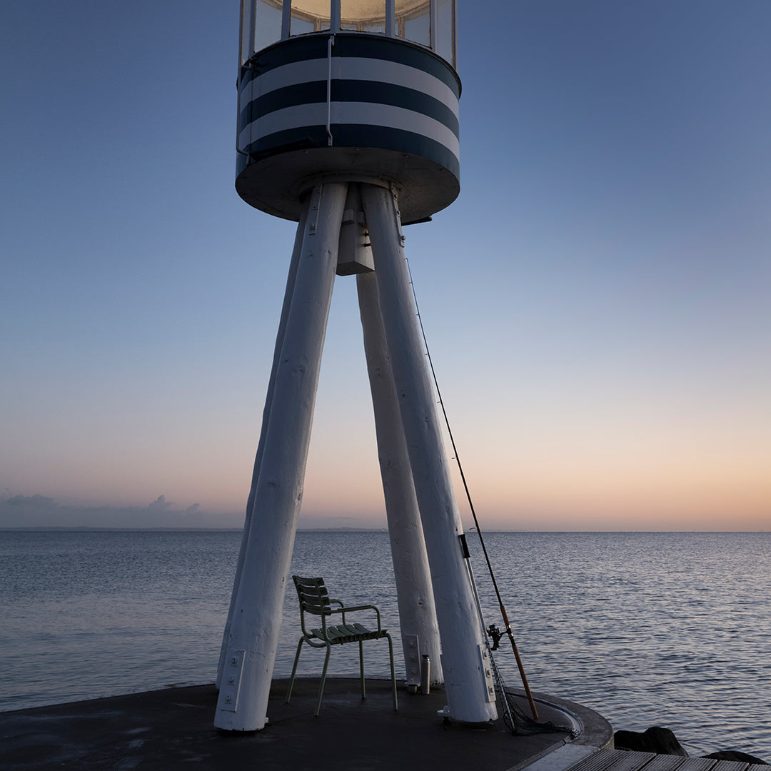 Lighthouse with blue and white stripes, white legs, set against a serene sunset over calm waters.